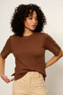 Woman wearing a brown sweater and beige pants against a plain background