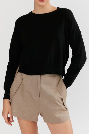 A model wearing a black long-sleeve crewneck sweater and beige shorts, cropped from the mouth down against a light gray background.