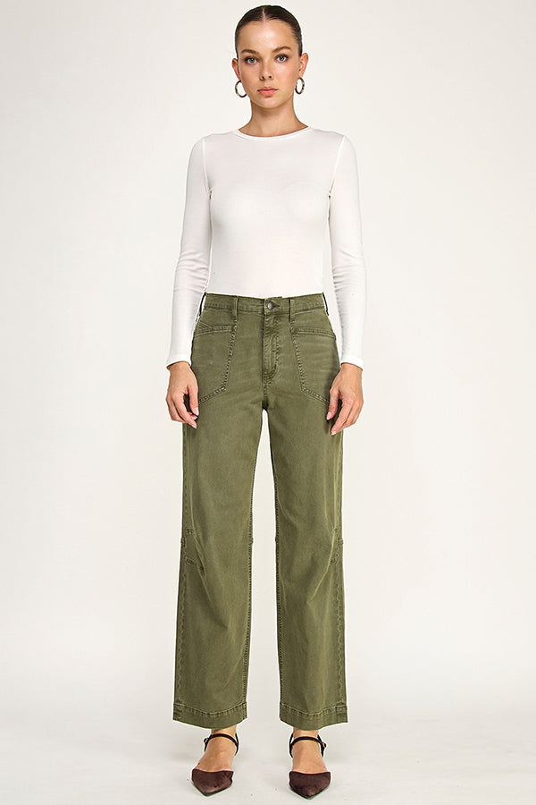 A model stands against a light gray studio background wearing a fitted white long-sleeve top, olive green wide-leg pants, and dark brown pointed-toe flats.