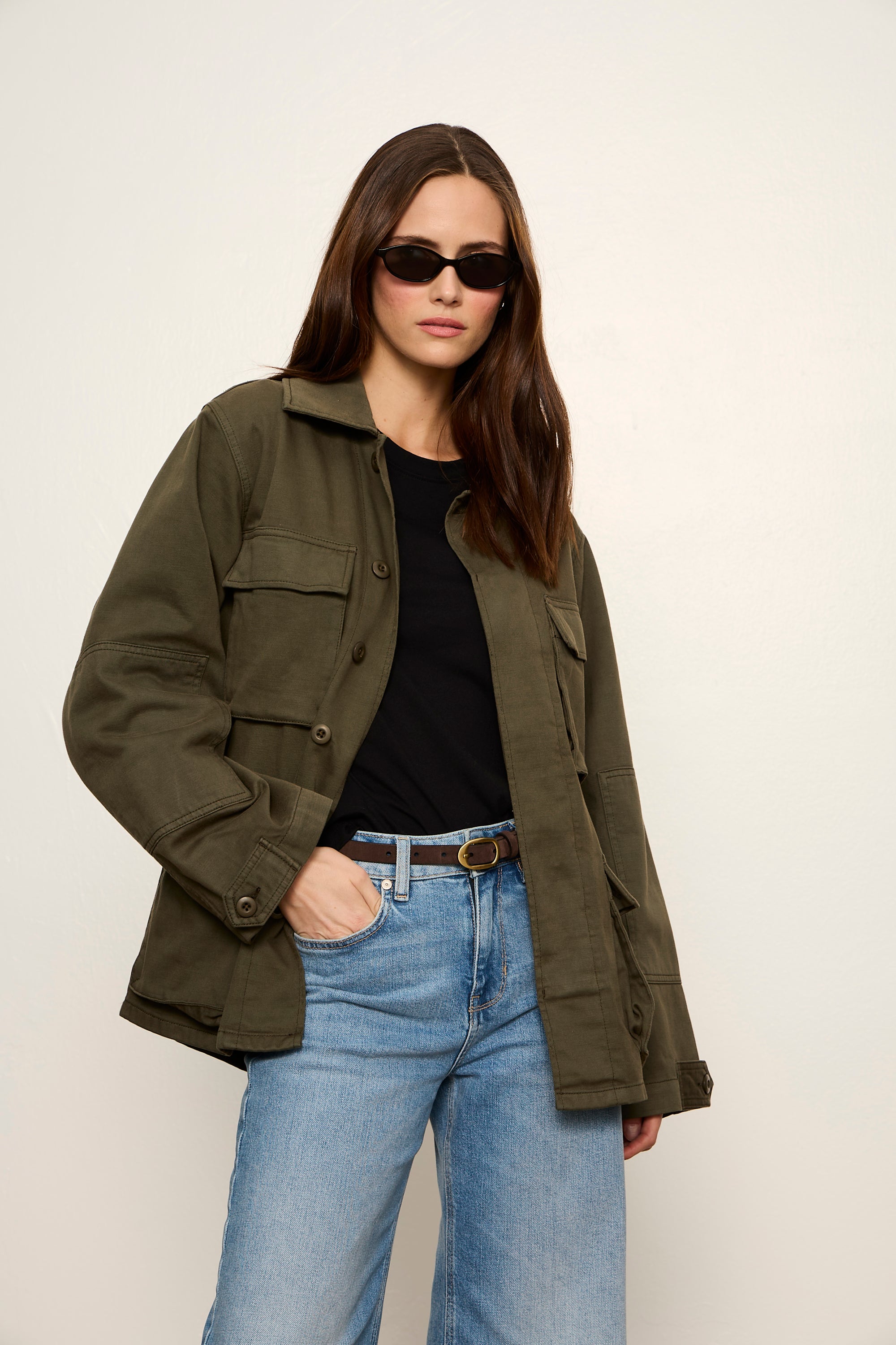 Person wearing an olive green jacket, black shirt, and blue jeans against a white background