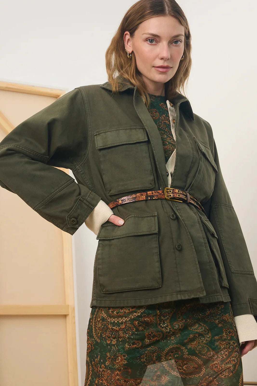 A woman models an olive green utility jacket with flap pockets and a matching belt, styled over a patterned green and brown dress against a neutral indoor background.