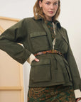 A woman models an olive green utility jacket with flap pockets and a matching belt, styled over a patterned green and brown dress against a neutral indoor background.