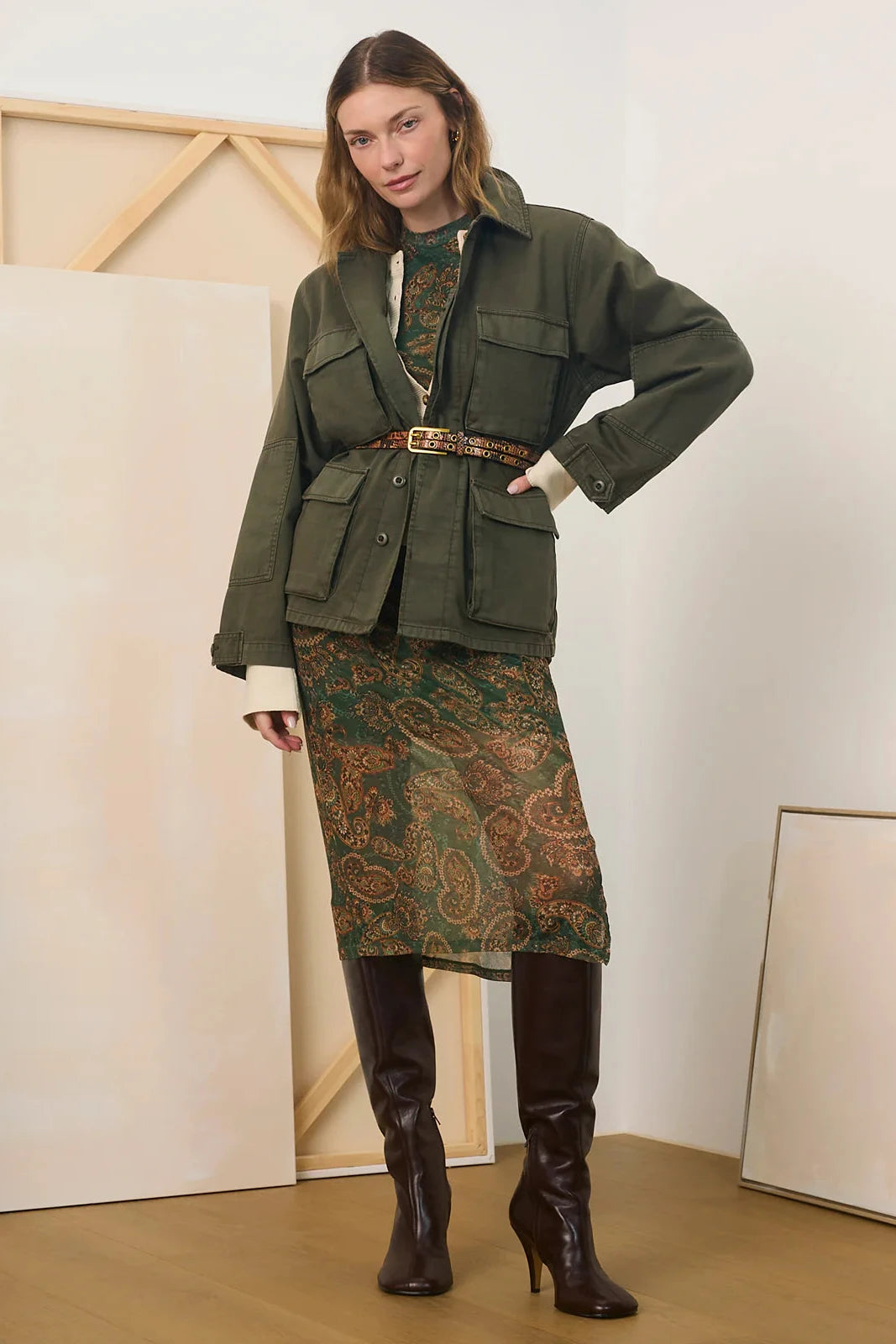 A model stands in a studio wearing an olive green utility jacket with a matching belt, a green paisley skirt, and dark brown knee-high boots.