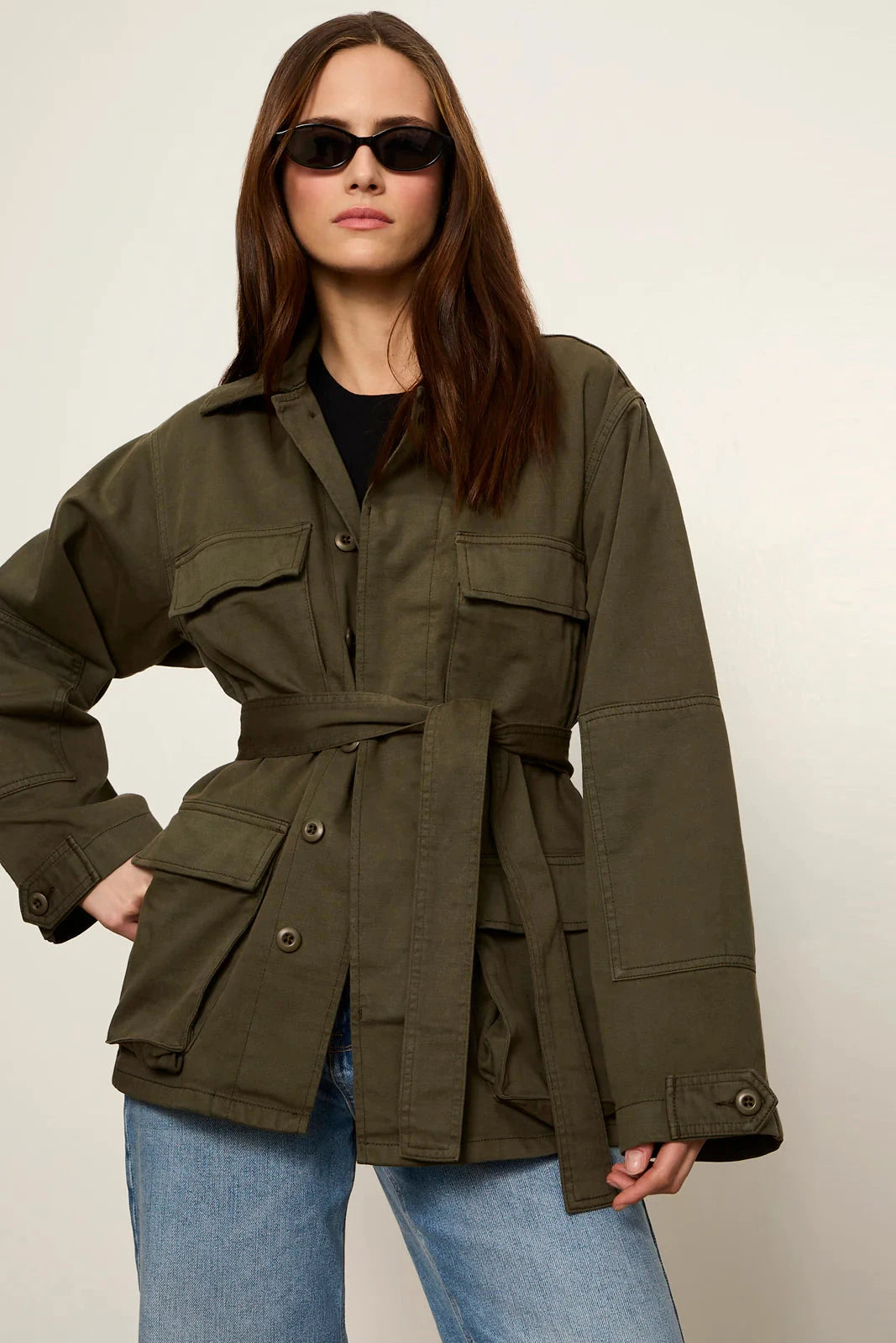 A woman modeling an oversized olive green utility jacket with a self-tie belt, worn over a black top and blue jeans against a plain light background.