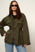 A woman modeling an oversized olive green utility jacket with a self-tie belt, worn over a black top and blue jeans against a plain light background.