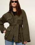 A woman modeling an oversized olive green utility jacket with a self-tie belt, worn over a black top and blue jeans against a plain light background.