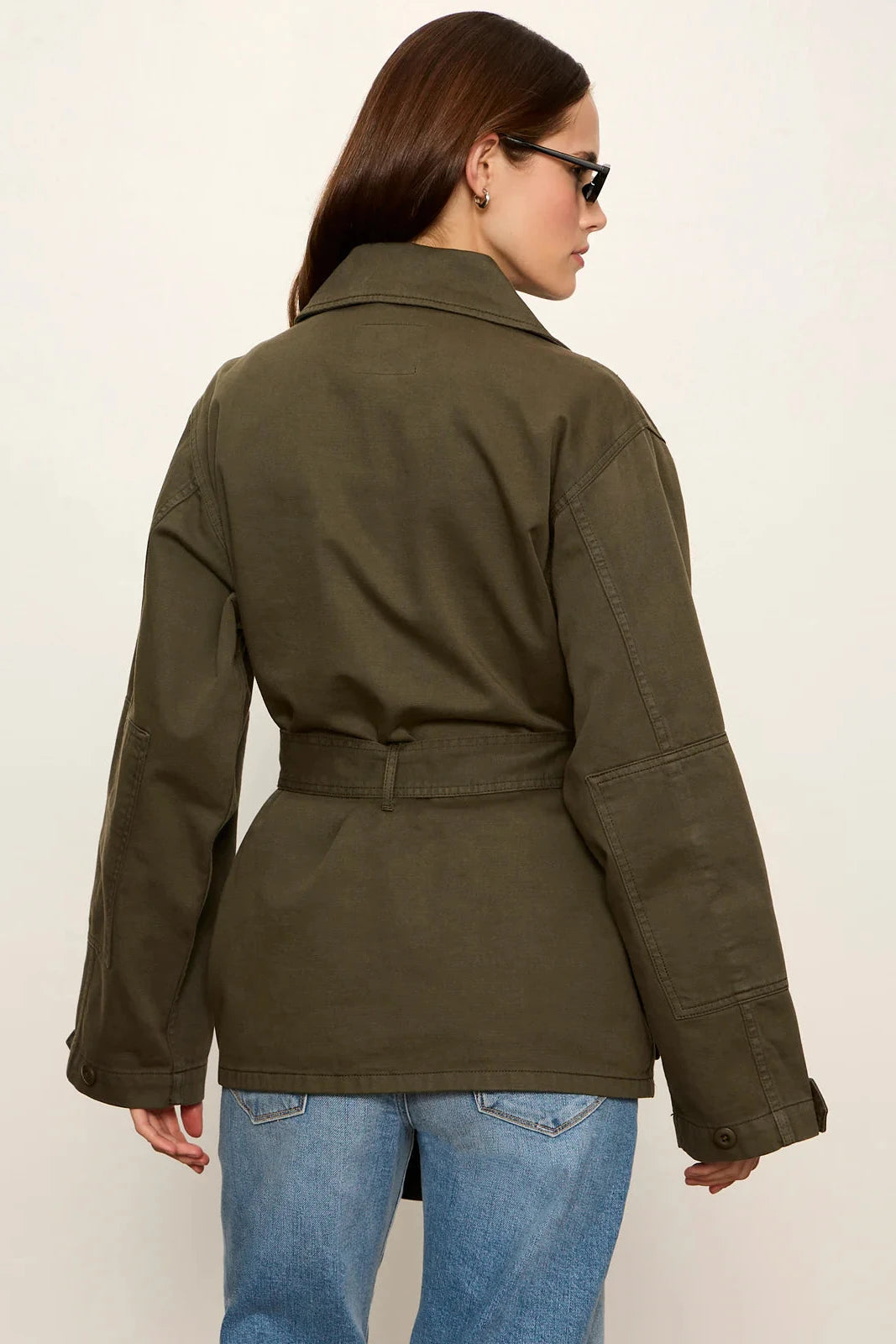 A woman shown from the back wearing an olive green belted utility jacket and blue jeans against a plain off-white studio background.