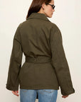 A woman shown from the back wearing an olive green belted utility jacket and blue jeans against a plain off-white studio background.