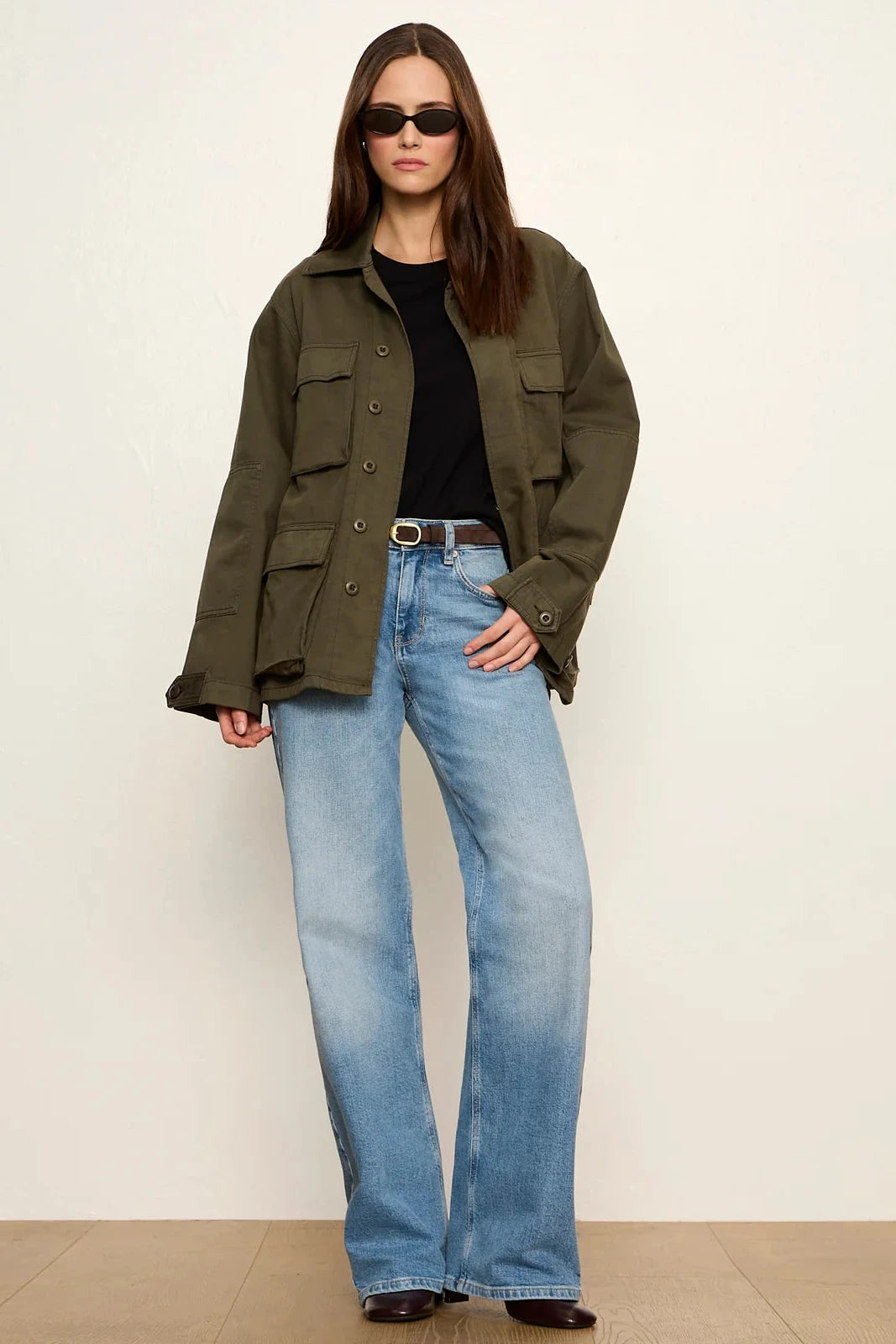 A woman modeling an olive green utility jacket over a black top and light blue wide-leg jeans, standing against a plain off-white wall.