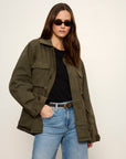 Person wearing an olive green jacket, black shirt, and blue jeans against a white background