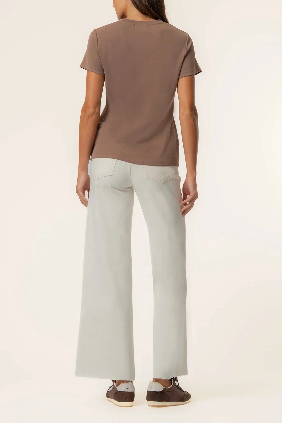 A woman shown from the back wearing a brown short-sleeve t-shirt, light gray wide-leg pants, and brown shoes against a plain off-white studio background.