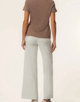 A woman shown from the back wearing a brown short-sleeve t-shirt, light gray wide-leg pants, and brown shoes against a plain off-white studio background.