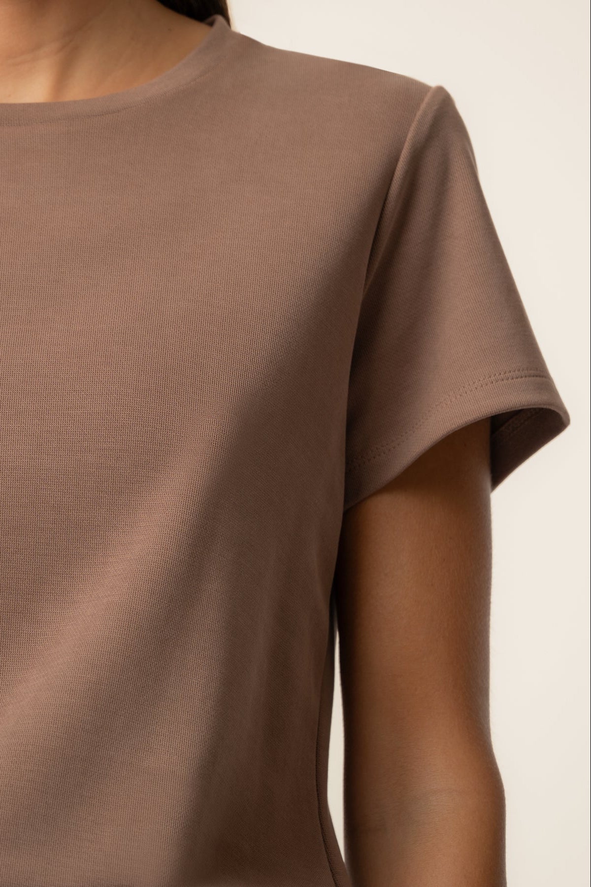 A close-up of a person wearing a plain short-sleeve crew-neck t-shirt in a muted taupe-brown color, set against a clean off-white background.