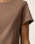 A close-up of a person wearing a plain short-sleeve crew-neck t-shirt in a muted taupe-brown color, set against a clean off-white background.