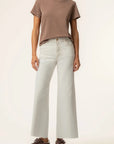 A model wearing a brown short-sleeve crewneck t-shirt, light gray wide-leg jeans, and dark brown lace-up sneakers, standing against a plain off-white studio background.