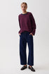 A woman modeling a dark purple and red striped long-sleeve knit top with navy blue wide-leg trousers and dark brown leather loafers, standing against a plain light gray studio background.