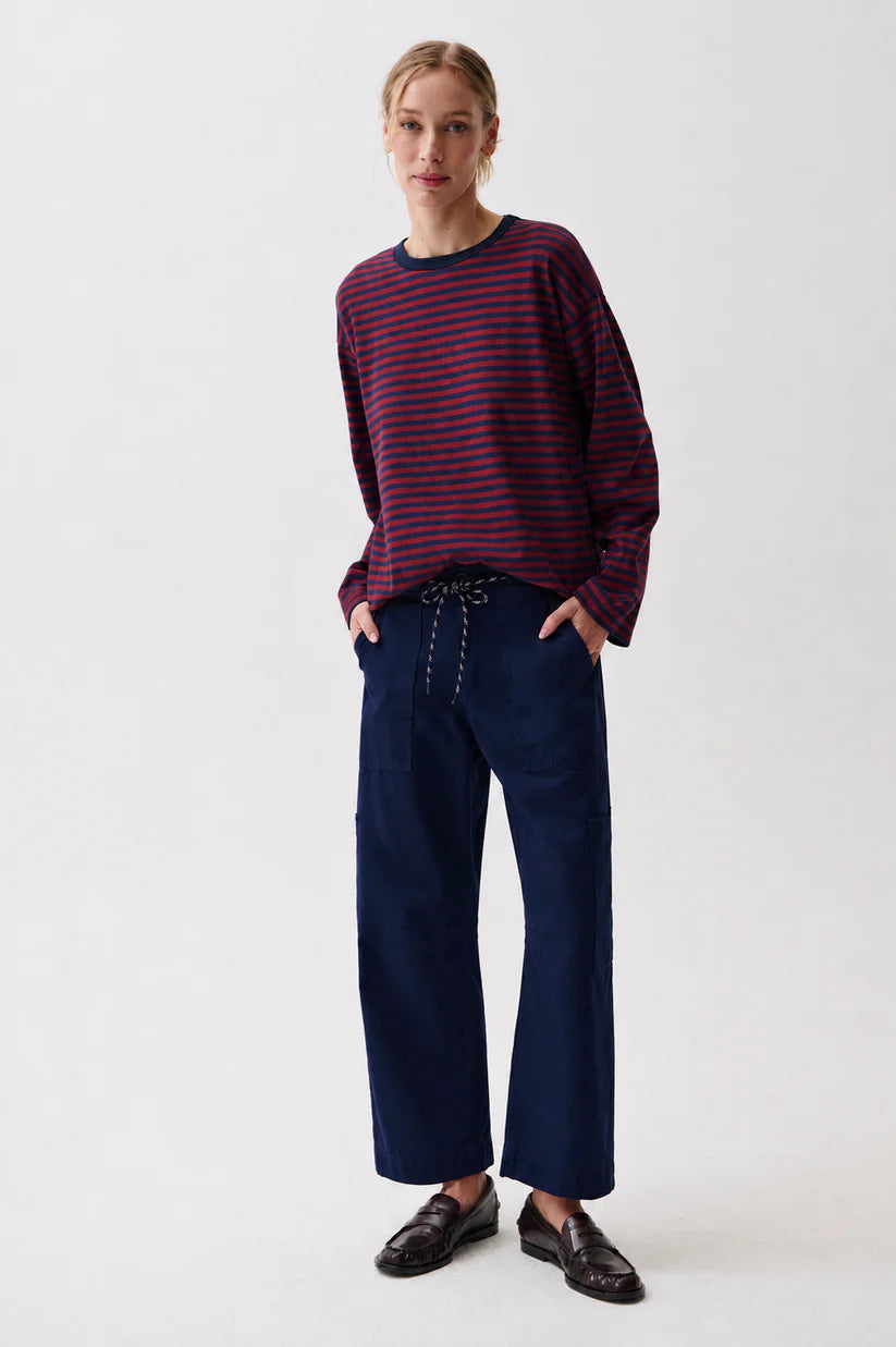A woman modeling a dark purple and red striped long-sleeve knit top with navy blue wide-leg trousers and dark brown leather loafers, standing against a plain light gray studio background.