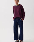 A woman modeling a dark purple and red striped long-sleeve knit top with navy blue wide-leg trousers and dark brown leather loafers, standing against a plain light gray studio background.