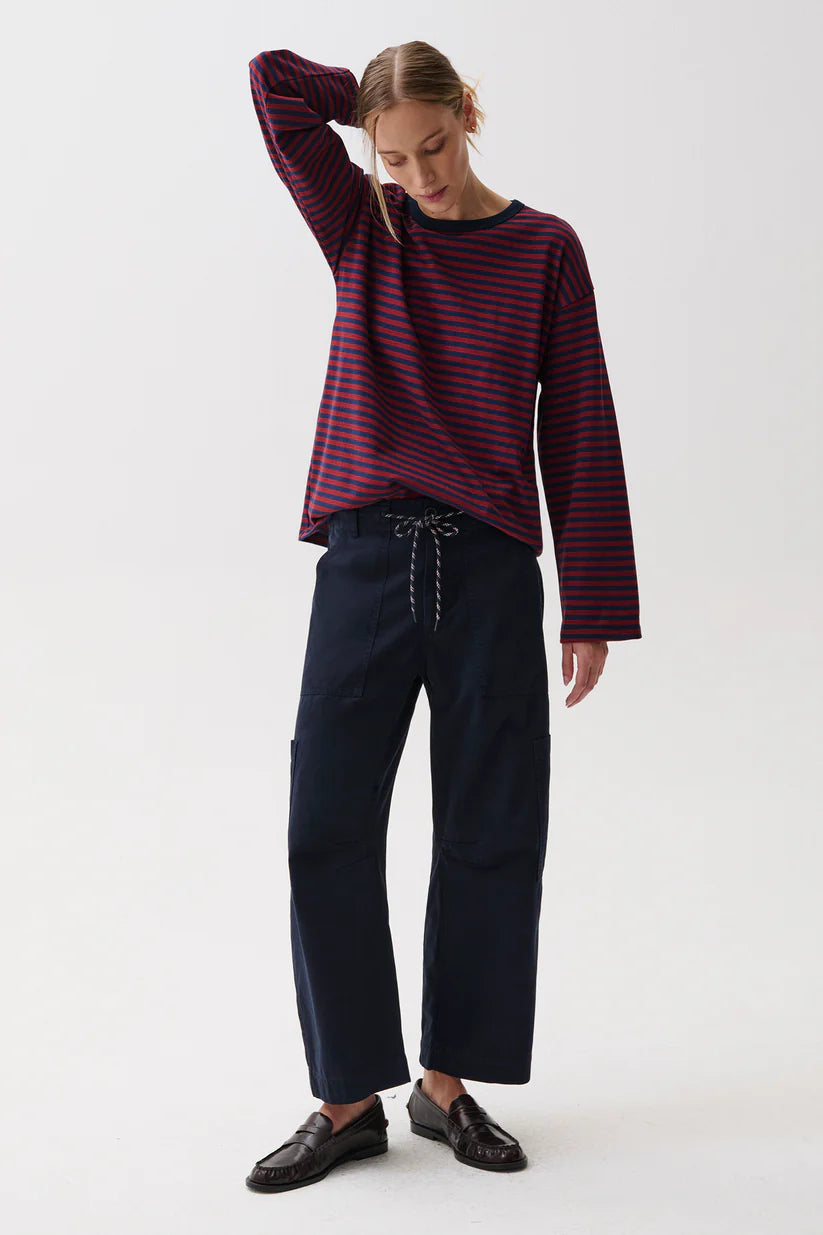 A woman modeling a dark red and navy blue horizontally striped long-sleeve top with dark navy wide-leg trousers and black leather loafers, standing against a plain white studio background.