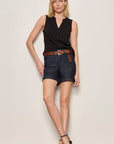 Woman wearing a black sleeveless top and dark shorts with a belt against a plain background
