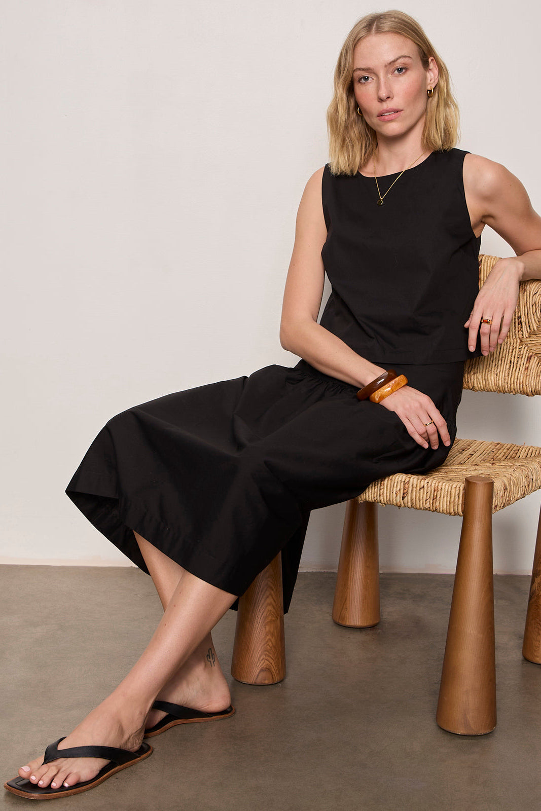 Woman in a black dress sitting on a wooden chair against a plain background