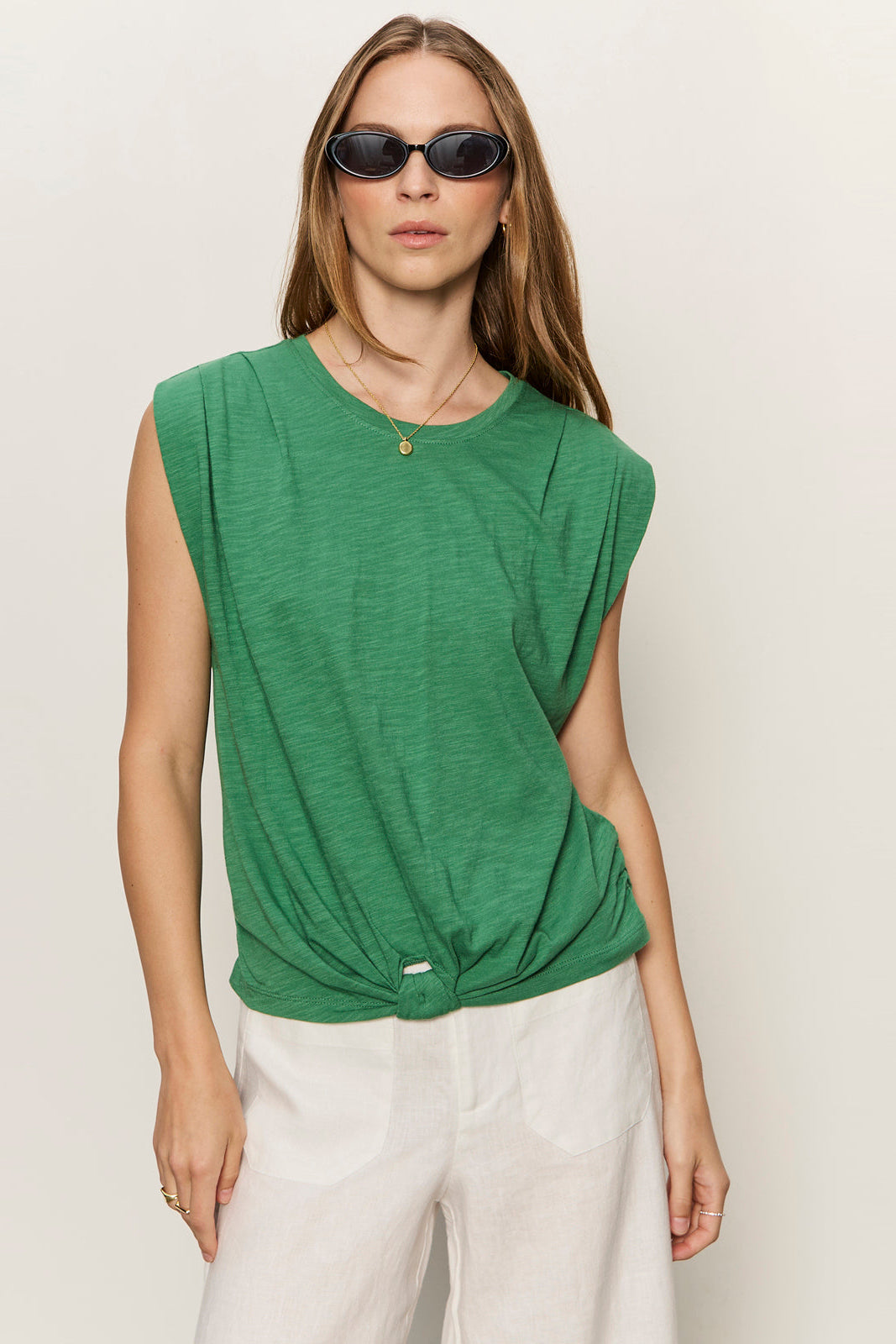 Woman wearing a green sleeveless top and white pants against a plain background