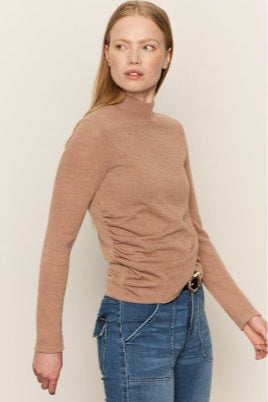 Woman wearing a brown turtleneck sweater and blue jeans against a beige background