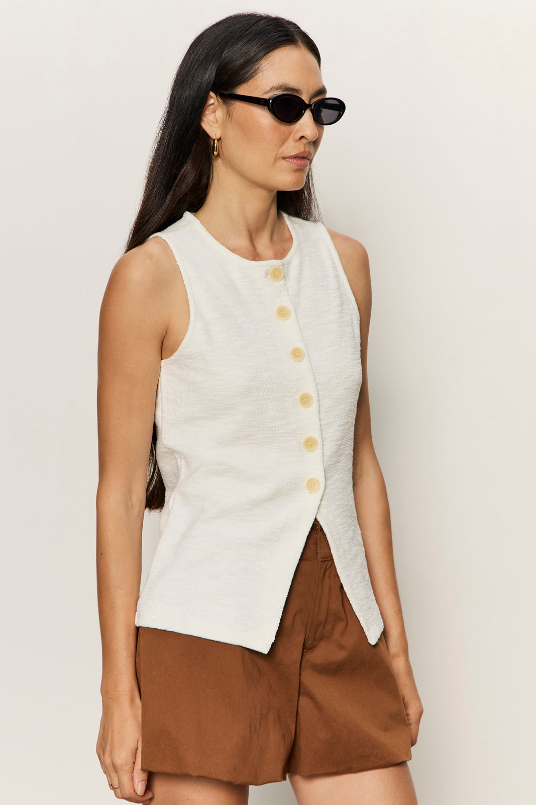 Woman wearing a white sleeveless button-up top and brown shorts against a plain background