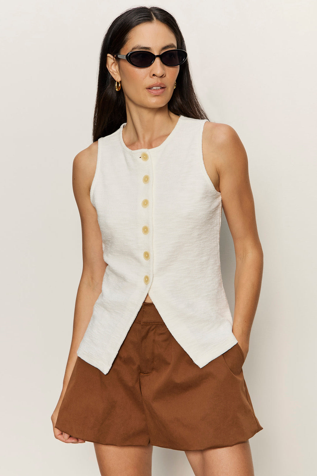 Woman wearing a white sleeveless vest with buttons and brown shorts against a plain background