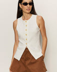 Woman wearing a white sleeveless vest with buttons and brown shorts against a plain background