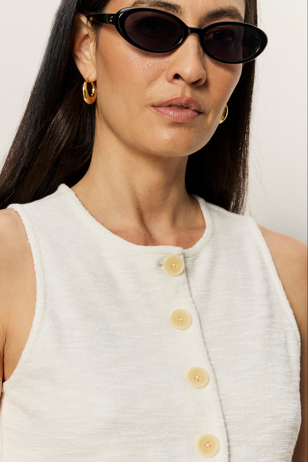 Woman wearing a white sleeveless top with yellow buttons and black sunglasses against a plain background