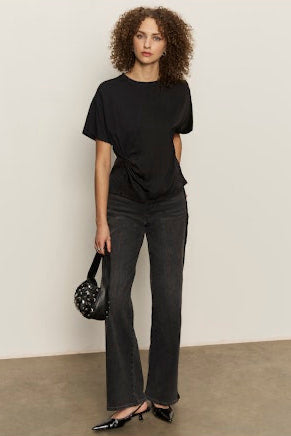 Woman wearing a black t-shirt and dark jeans holding a black handbag against a plain background