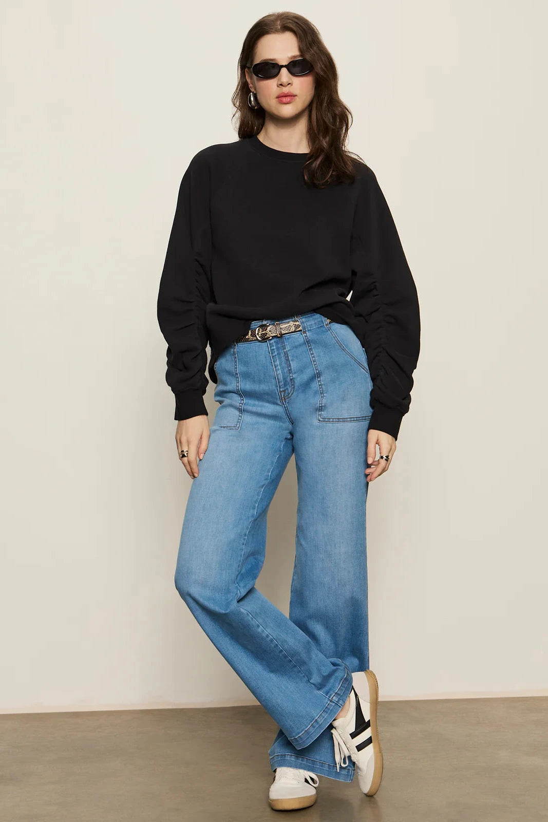 Woman wearing a black sweater, blue jeans, and sunglasses against a plain background