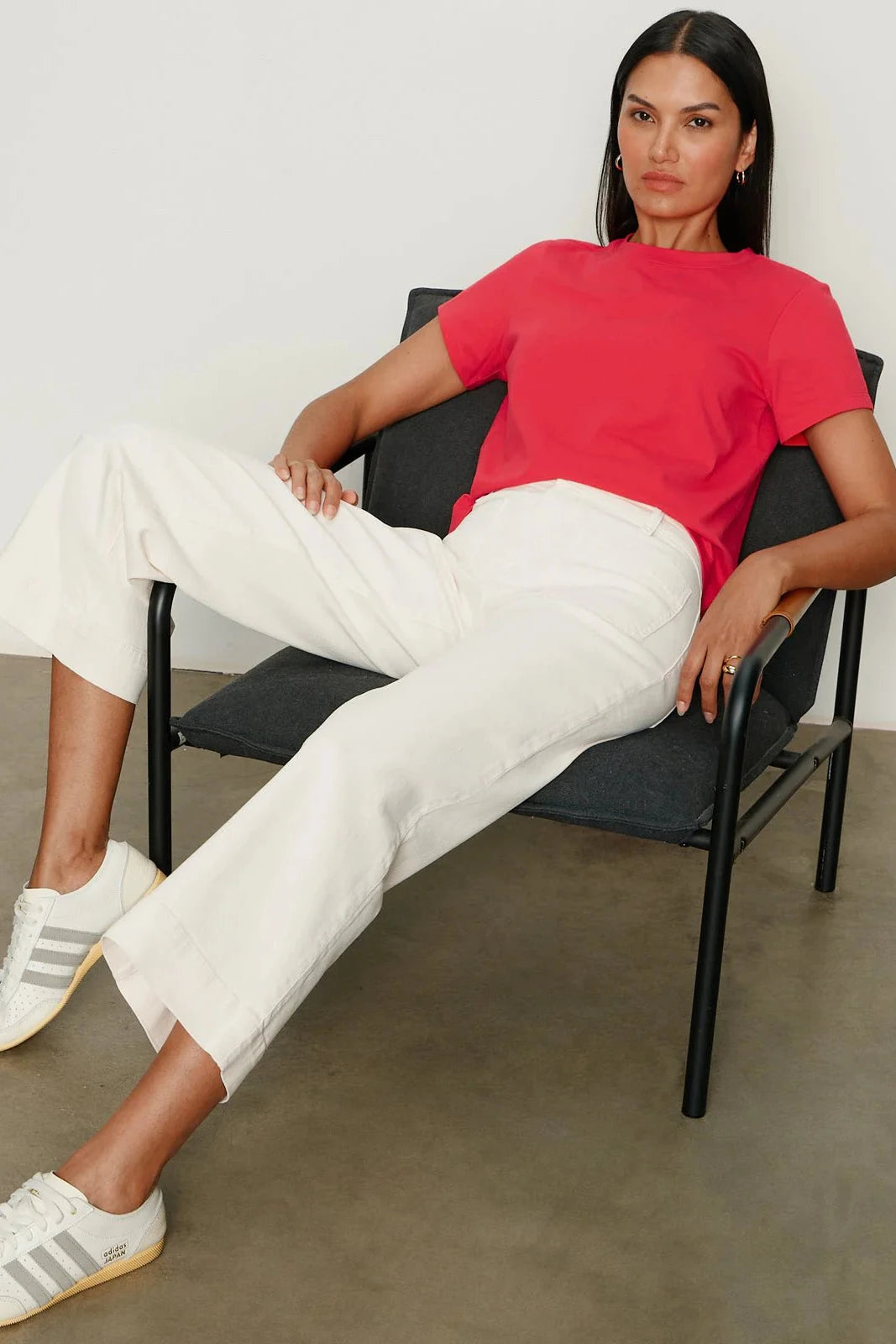 Woman sitting on a chair wearing a red shirt and white pants with Adidas shoes.