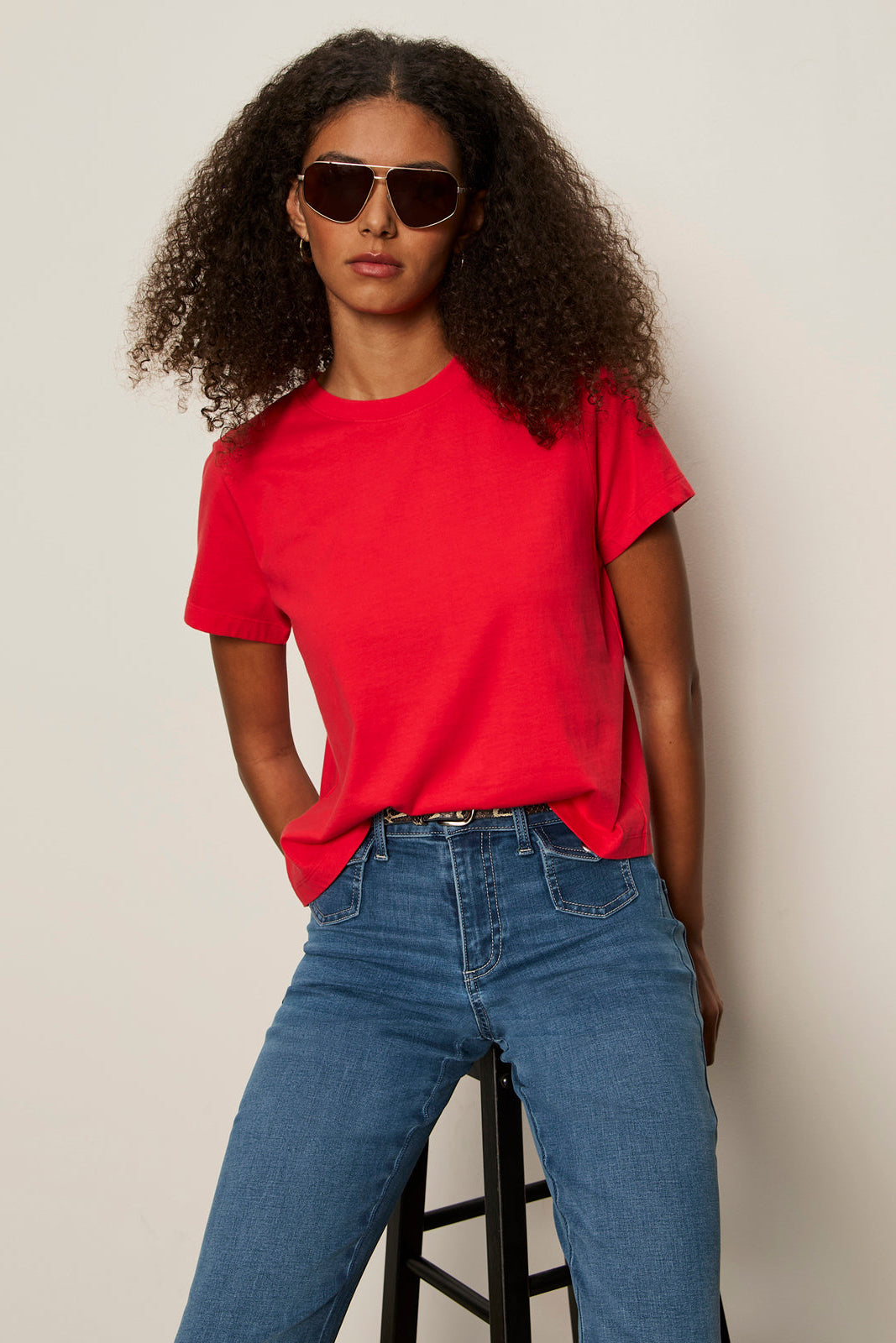 Person wearing a red t-shirt and blue jeans with sunglasses on a plain background