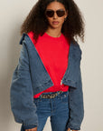 Woman wearing a red shirt, blue denim jacket, and jeans with sunglasses on a beige background
