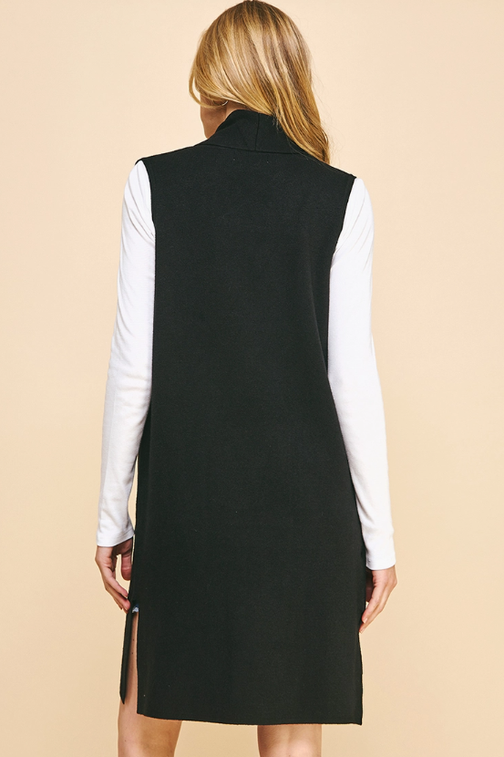 A woman shown from the back wearing a black sleeveless mock-neck longline vest over a white long-sleeve shirt, set against a solid beige background.
