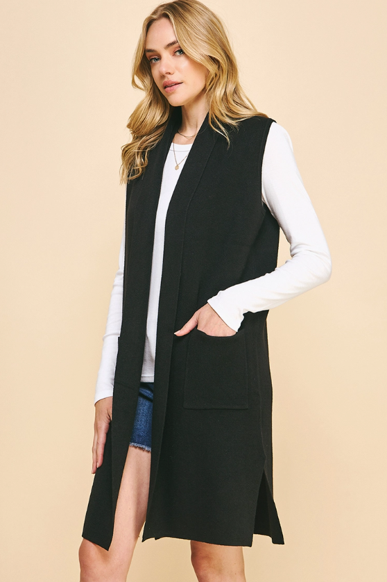 A woman modeling a long black sleeveless vest over a white long-sleeve top and denim shorts, standing against a solid beige studio background.