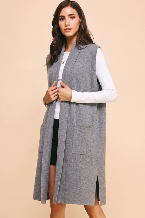 A woman modeling a long gray sleeveless knit vest over a white long-sleeve top and black skirt, standing against a solid beige studio background.