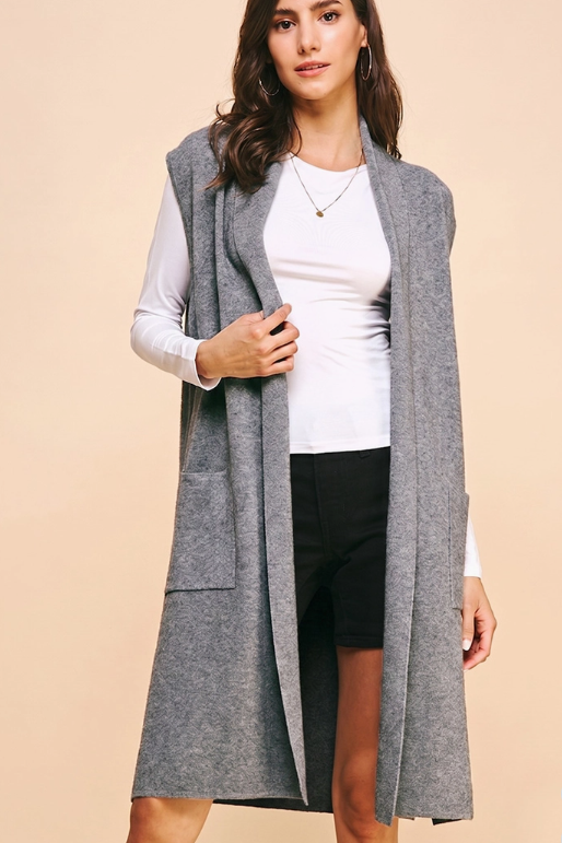 A woman models a long, sleeveless gray knit duster vest over a white long-sleeve top and black shorts, set against a solid beige studio background.