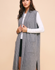 A woman modeling a long gray sleeveless knit vest over a white long-sleeve top and black skirt, standing against a solid beige studio background.