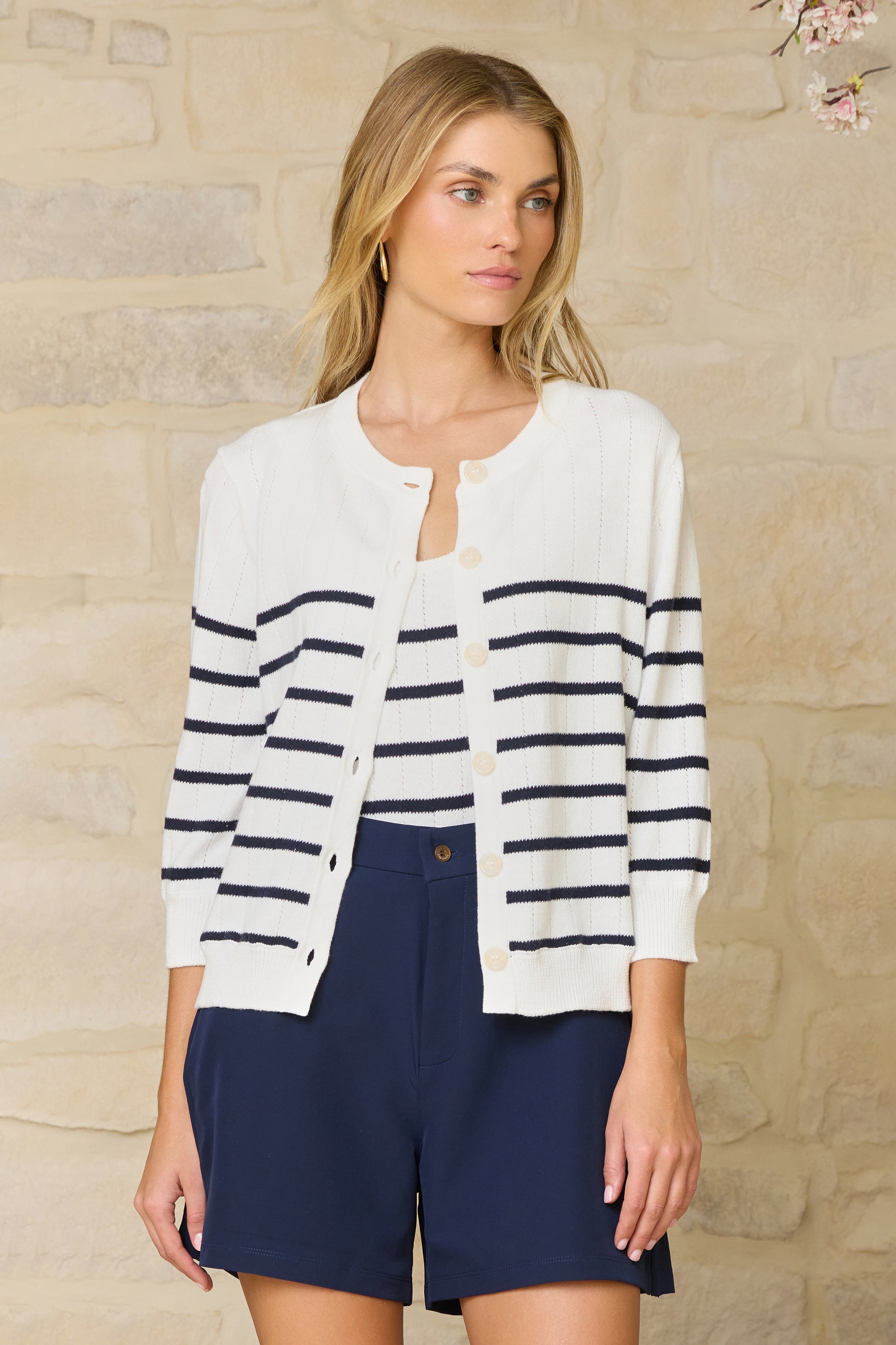 Woman wearing a white cardigan with navy stripes and navy shorts against a stone wall.