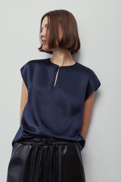 A woman wearing a navy blue short-sleeve top with a small gold-toned zipper detail at the neckline, paired with black satin-like pants, photographed against a plain light gray background.
