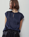 A woman wearing a navy blue short-sleeve top with a small gold-toned zipper detail at the neckline, paired with black satin-like pants, photographed against a plain light gray background.