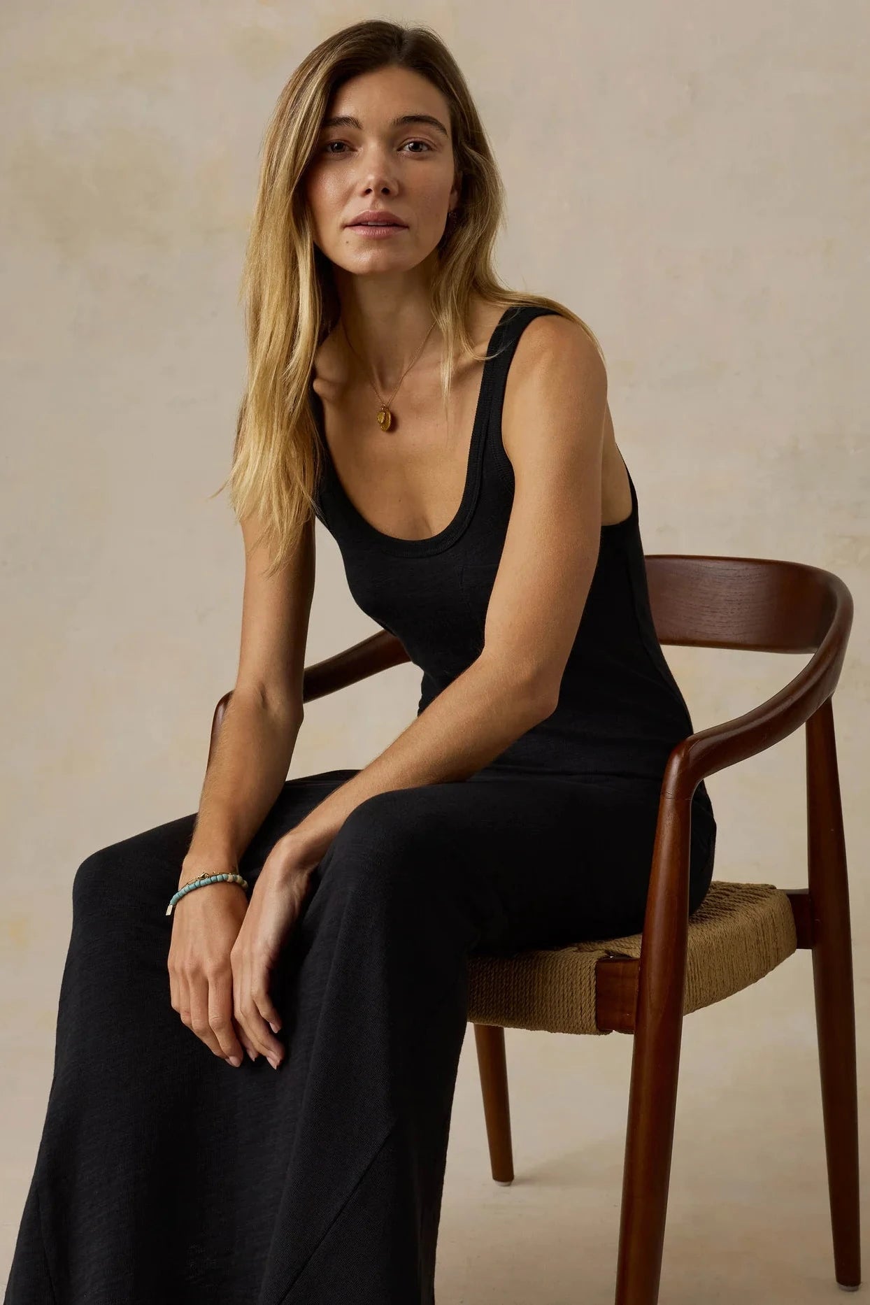 Woman in a black sleeveless dress sitting on a wooden chair against a beige background