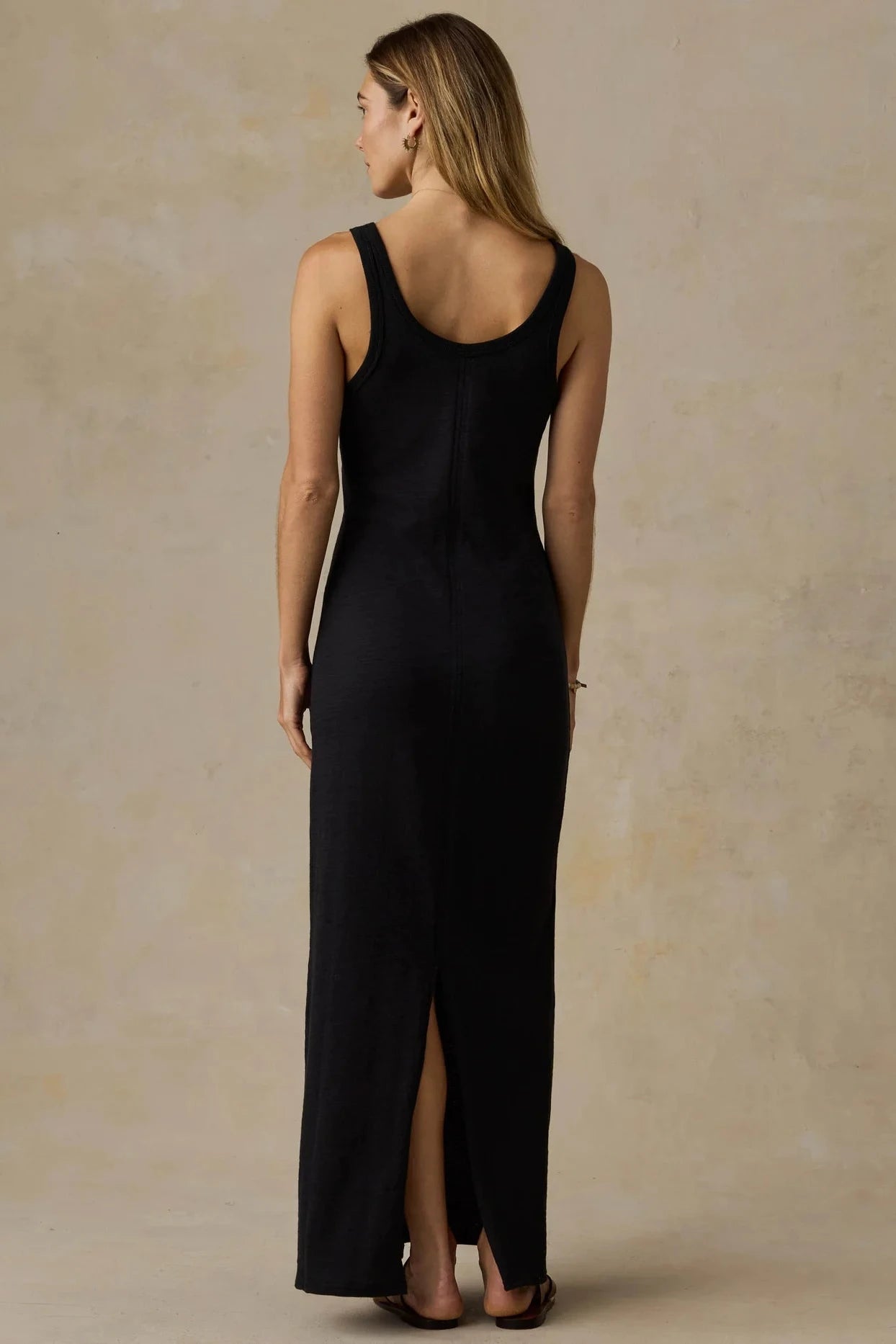 A woman wearing a sleeveless black maxi dress with a high front slit, shown from the back against a textured beige studio backdrop.