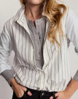 A woman wearing a light gray and white vertically striped button-up shirt layered over a gray henley tee, paired with black jeans.