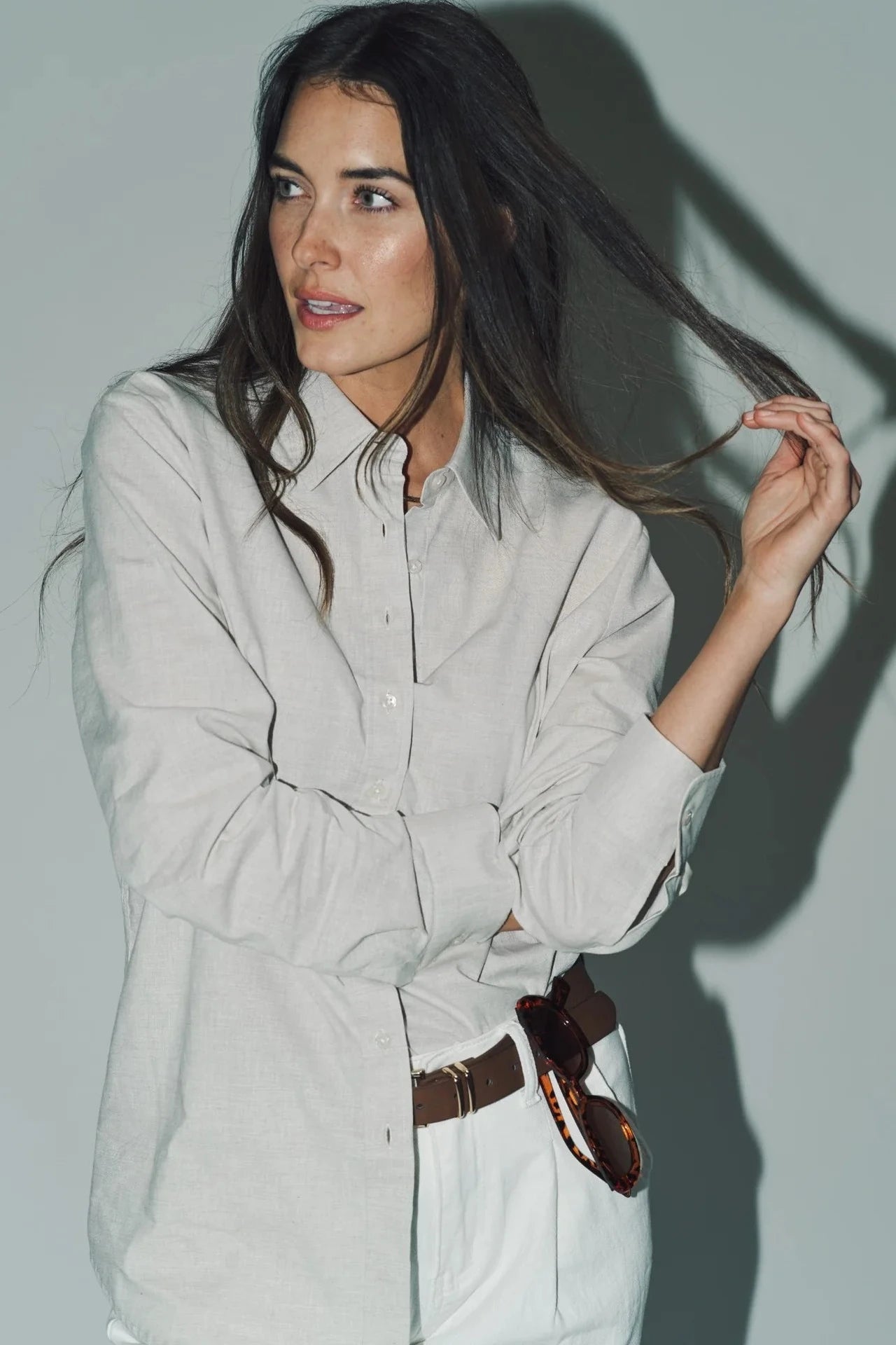 Woman wearing a light gray shirt with rolled-up sleeves, holding her hair against a plain background