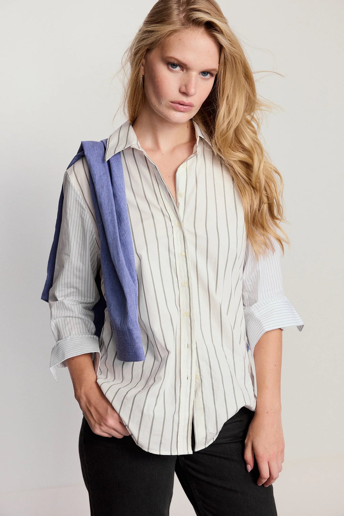 Woman wearing a white striped shirt with rolled-up sleeves, holding a blue towel over her shoulder against a plain background.