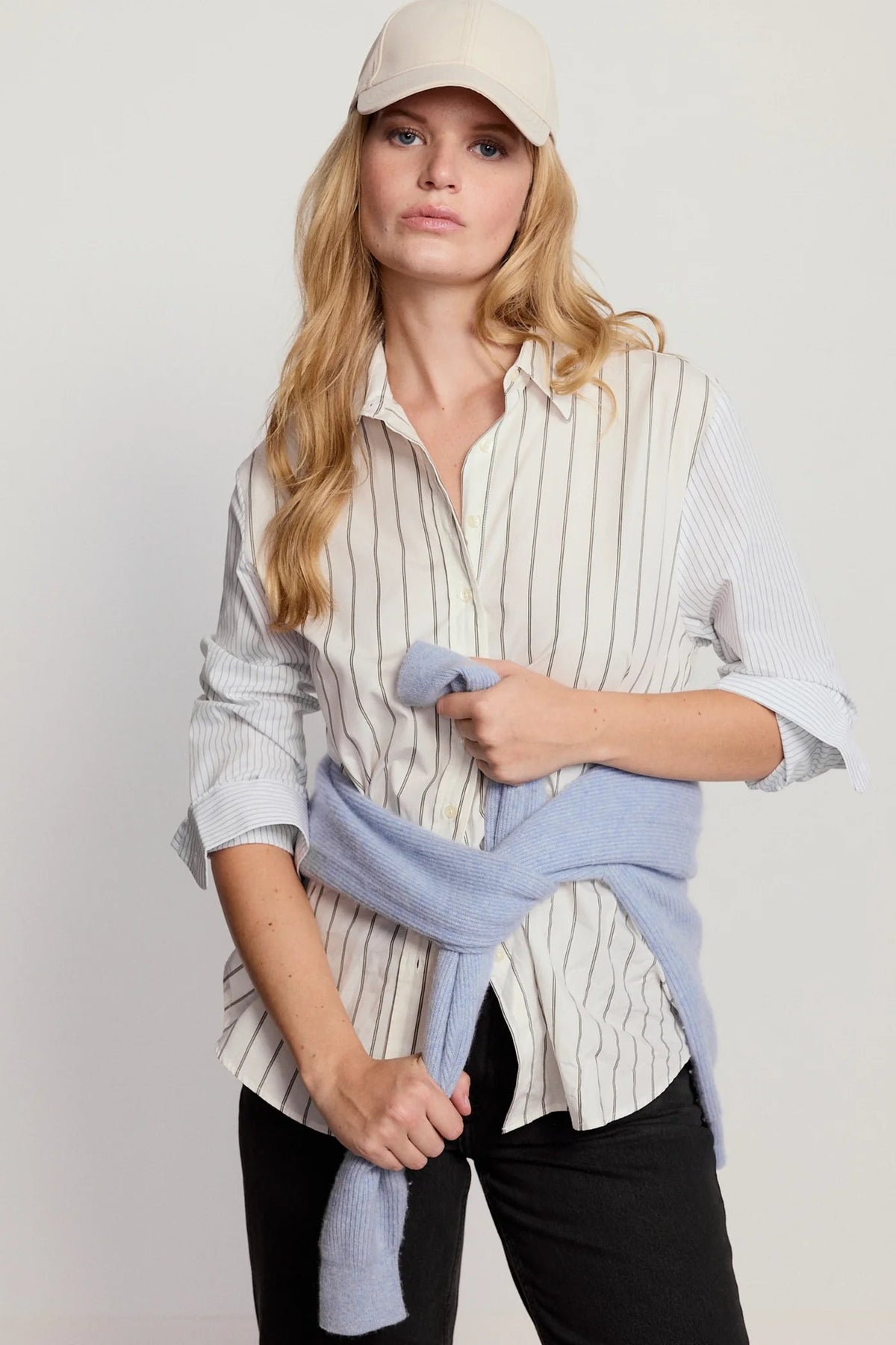 A woman modeling a light beige baseball cap, a cream and navy vertically striped button-down shirt, and a light blue knit sweater tied around her waist, paired with black pants, against a plain light gray background.
